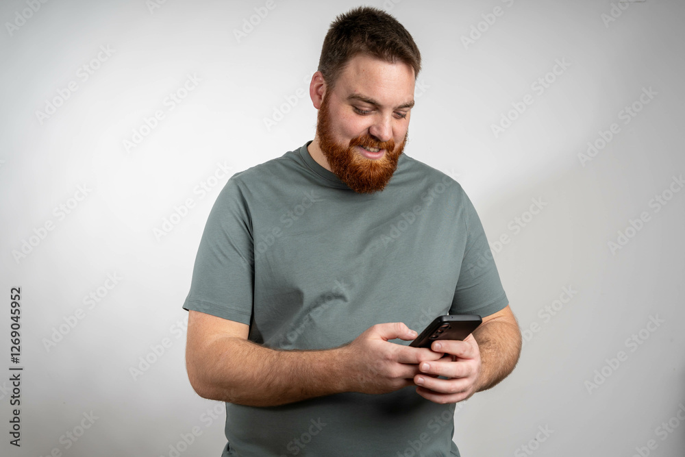 A man with a beard is holding a cell phone in his hand, looking at the screen with a focused expression. He appears to be typing or scrolling on the device. A man with a beard is holding a cell phone in his hand, looking at the screen with a focused expression. He appears to be typing or scrolling on the device.