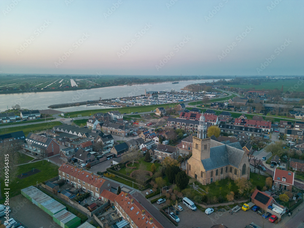 Fototapeta premium Aerial view of a Dutch Catholic church on the banks of the De Lek river in the southern Netherlands. Spring landscape with houses clustered on the vast plain of the Netherlands in the evening at sunse