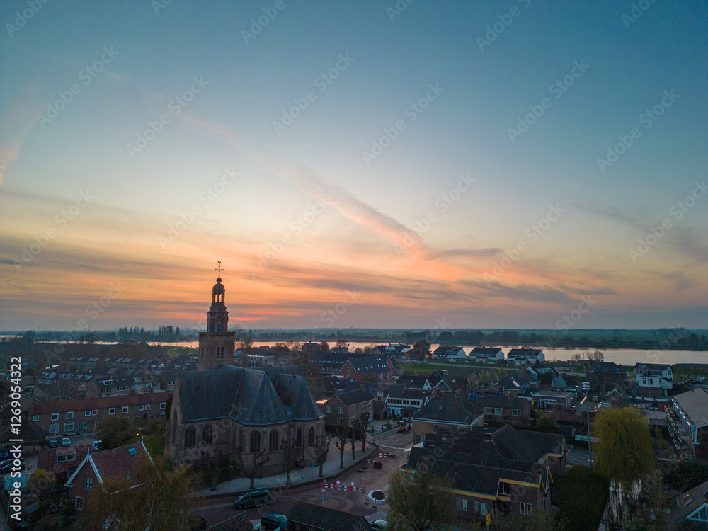 Fototapeta premium Aerial view of a Dutch Catholic church on the banks of the De Lek river in the southern Netherlands. Spring landscape with houses clustered on the vast plain of the Netherlands in the evening at sunse