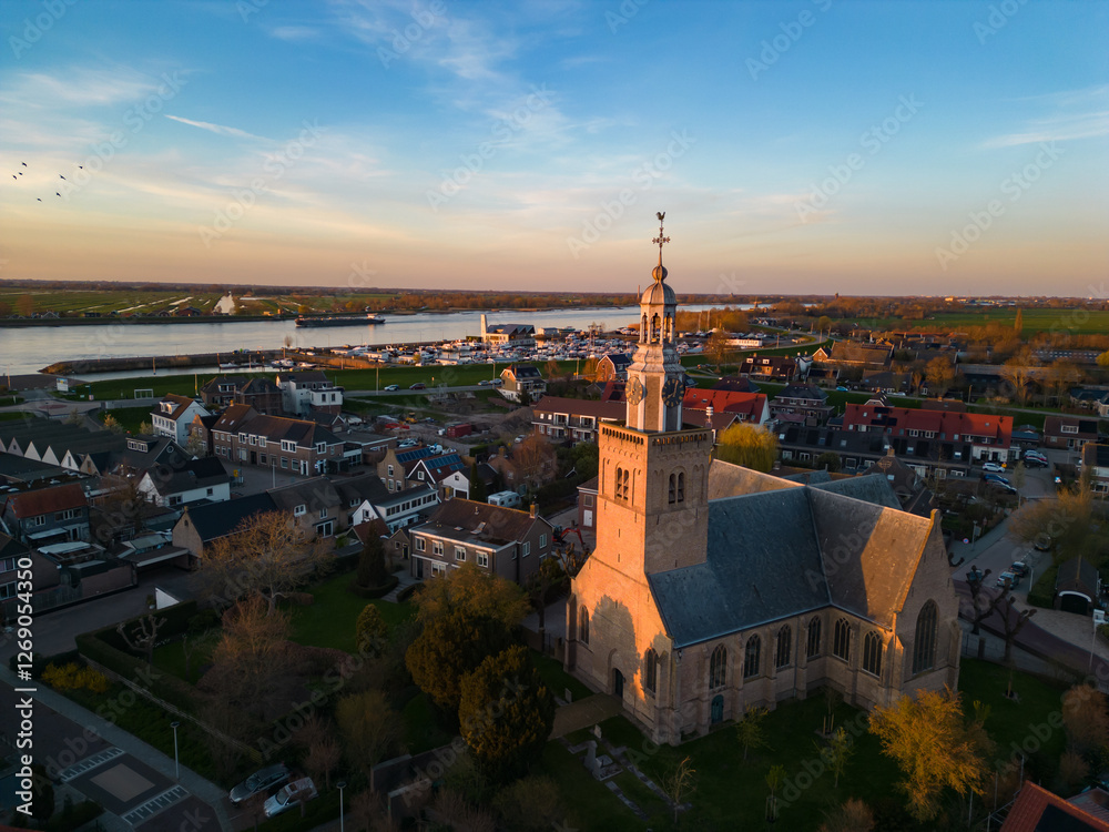 Fototapeta premium Aerial view of a Dutch Catholic church on the banks of the De Lek river in the southern Netherlands. Spring landscape with houses clustered on the vast plain of the Netherlands in the evening at sunse