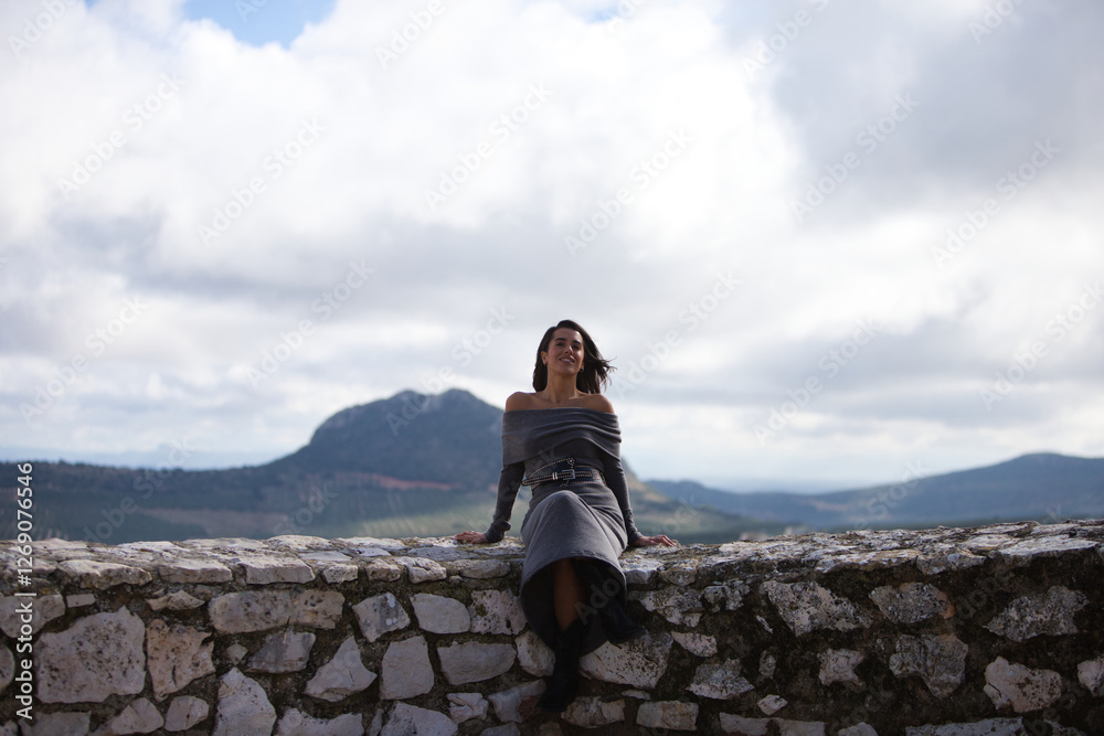 Young and beautiful woman, brunette with short hair, dressed in gray with black boots and leather belt sitting on the stone wall, in the background the mountains with Indian face. Young and beautiful woman, brunette with short hair, dressed in gray with black boots and leather belt sitting on the stone wall, in the background the mountains with Indian face.