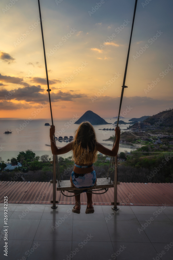 A peaceful scene shows a person swinging joyfully at sunset over a shimmering ocean, with a distant tropical island, offering an escape from daily life, Komodo, Indonesia