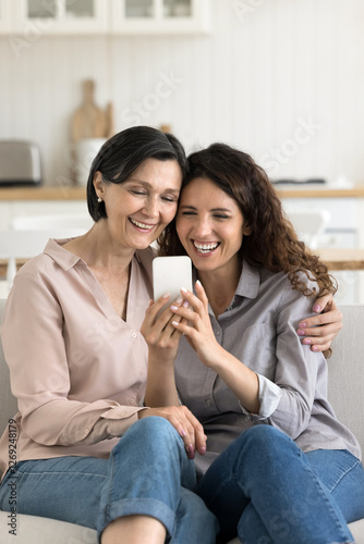 Family leisure time and digital tech. Laughing candid young daughter elderly mom cuddle on sofa at kitchen relax shoot funny selfie photo on mobile phone enjoy hilarious vlogs read funny message text