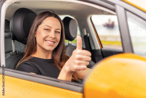 A young woman with long brown hair and a bright smile sits in the driver's seat of a yellow car, giving a thumbs-up gesture through the open window with a confident and positive expression.