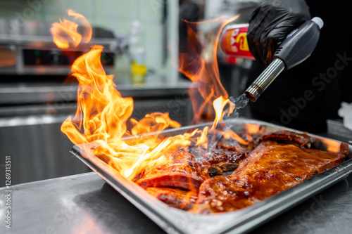 A chef using a culinary torch to flambé a tray of marinated meat, creating dramatic flames in a busy kitchen environment