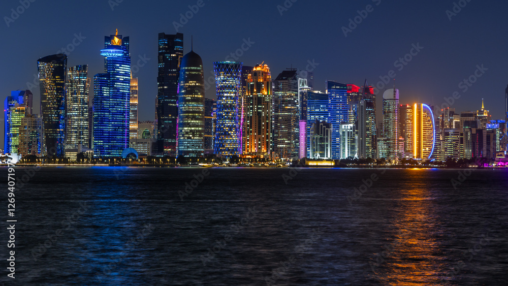 Naklejka premium Doha city business downtown center panorama illuminated at night, with modern skyscrapers and sea in the foreground, Qatar