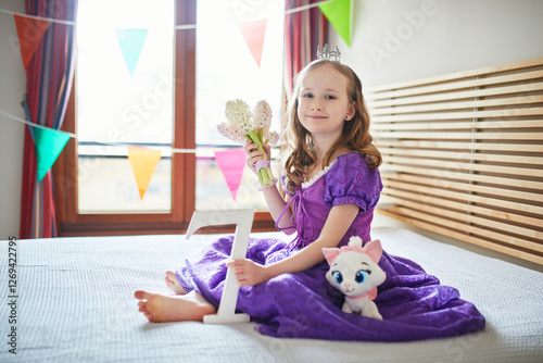 Adorable preschooler girl in red dress sitting on bed with white hyacinths and white wooden number seven. Seventh birthday concept. Kid celebrating seventh birthday.