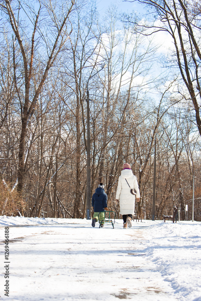 Kyiv, Ukraine - 02 16 2025: Unknown mother and son with sled walking in winter park. Boy pulling sled with mom in snowy park. Winter activity. Family lifestyle. Toddler with mom from the back