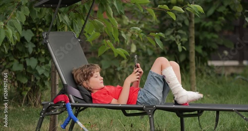 Little boy with fractured leg in gypsum plaster cast lying down on garden lounger near his medical orthopedic crutches, relaxing by playing a game on his mobile phone