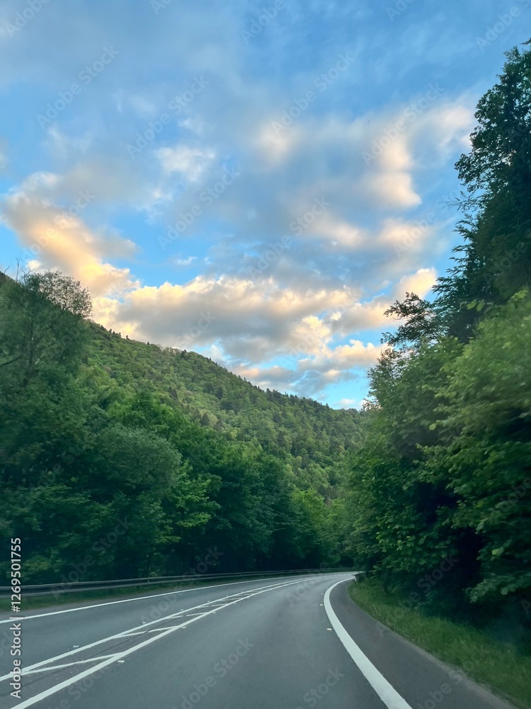 Fototapeta premium A winding road through lush green mountains under a blue sky with golden clouds, peaceful and scenic.