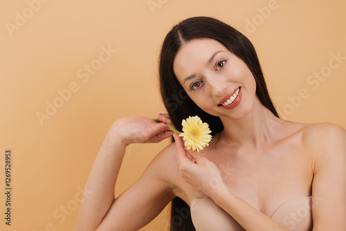 Tableau sur toile Beautiful smiling woman holding yellow gerbera flower near face on a beige background