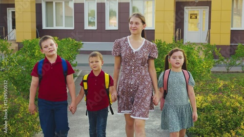 Mother leading children from school, female mother holding children hand, happy family, young nanny with children, schoolchildren with backpacks walking with aunt by the hand, little boys girl walking