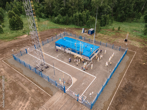 Aerial view of remote industrial facility with blue roof and fenced perimeter in forested area.