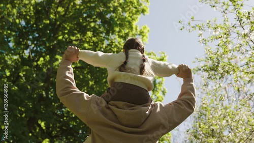 A man is holding a child on his shoulders in a park. The child is wearing a white sweater. The scene is peaceful and serene, with the man and child enjoying the outdoors together