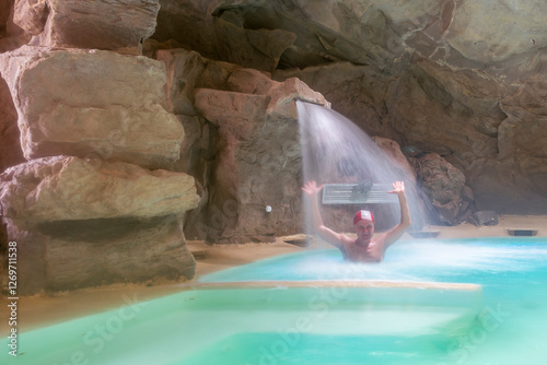 A man wearing a white and red cap under a waterfall in a spa inside a cave, Tuscany, Italy photo