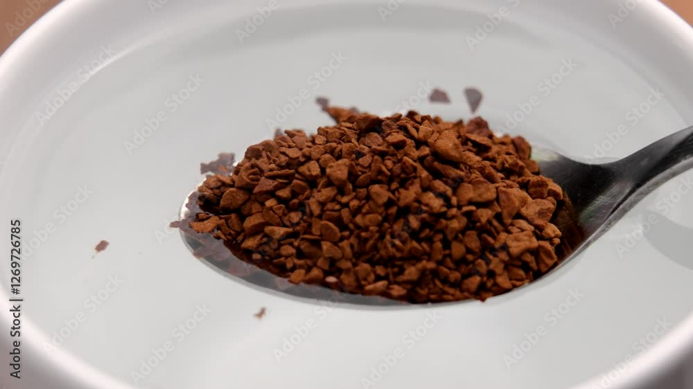 Metal spoon with instant granulated coffee sinks into a white cup of hot water, dissolving aromatic granules Metal spoon with instant granulated coffee sinks into a white cup of hot water, dissolving aromatic granules