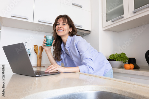 Beautiful curly brunette holding the glass of water, smiling and working at the laptop in the kitchen at home