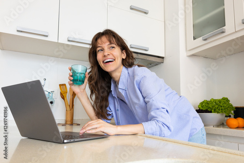 Beautiful curly brunette holding the glass of water, smiling and working at the laptop in the kitchen at home
