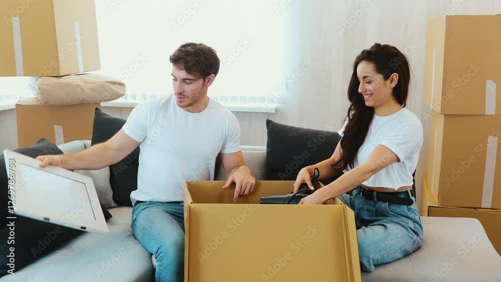 Young couple move into new apartment. Camera moves to right where man and woman sitting and unpacking box with stuff. Take out frame, hair dryer and a roll. Young couple move into new apartment. Camera moves to right where man and woman sitting and unpacking box with stuff. Take out frame, hair dryer and a roll.