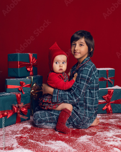 Obraz na płótnie brother and sister are sitting on the floor among green New Year gift boxes on a red background with snow. w salonie
