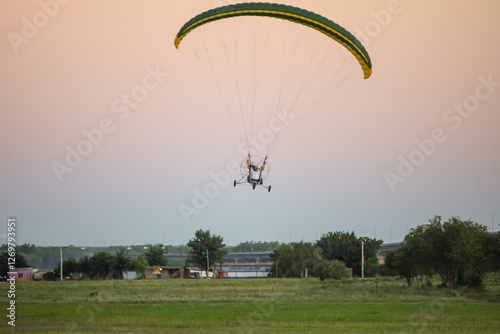 A paraglider soars through the sky above the mountains, carried by the wind. The paraglider is a bright orange color, and the pilot is silhouetted against the sky. The mountains are green and lush, an