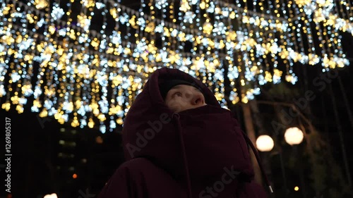 A girl in a hood in winter looks at the light above the street at night.