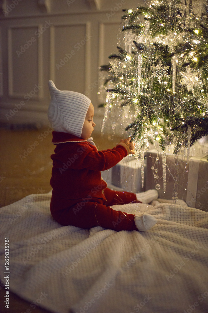 One little girl in a red suit sits on a white carpet at the Christmas tree at home for 1 year.