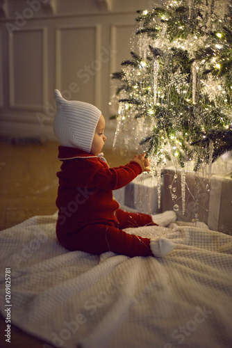 Obraz na płótnie One little girl in a red suit sits on a white carpet at the Christmas tree at home for 1 year. w salonie