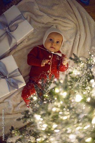 Obraz na płótnie One little girl in a red suit sits on a white carpet at the Christmas tree at home for 1 year. w salonie
