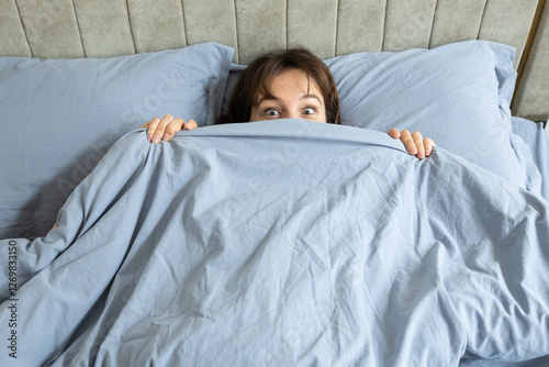Beautiful playful brunette looking out surpisedly of the blanket in the bed at home in the morning