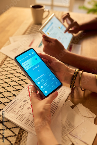 Hands holding smartphone and tablet while managing personal finances and analyzing expenses through apps, with documents on table and coffee in background
