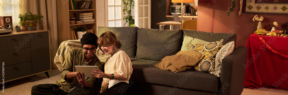Happy couple sitting on floor next to couch with large tablet in hands, smiling. Warm and cozy atmosphere with bookshelves and decorations in background Happy couple sitting on floor next to couch with large tablet in hands, smiling. Warm and cozy atmosphere with bookshelves and decorations in background