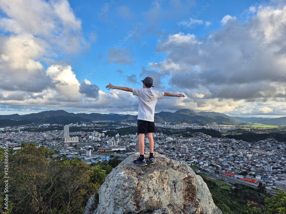 Obraz premium menino no alto da montanha com braços abertos observando a vista da cidade 