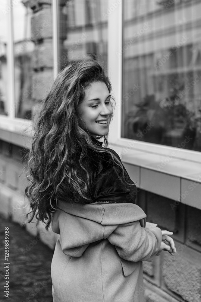 Fototapeta premium woman with long wavy hair. She is wearing a coat and stands sideways to the camera, turning her head slightly back and smiling. In the background you can see the wall of a building with large windows.