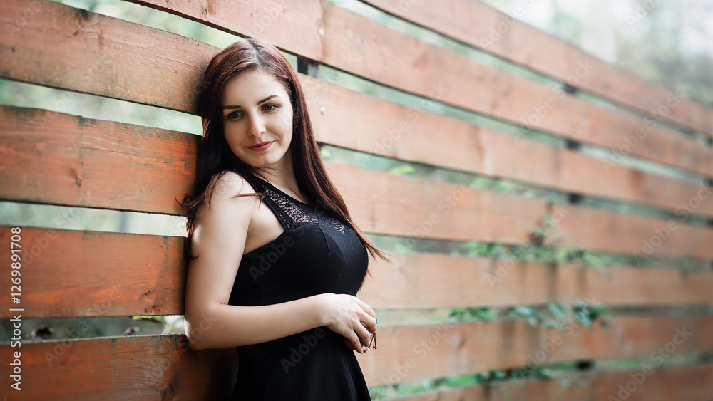 A woman stands with her back slightly leaning against a wooden fence made of horizontal boards. She looks away with a slight smile on her face.