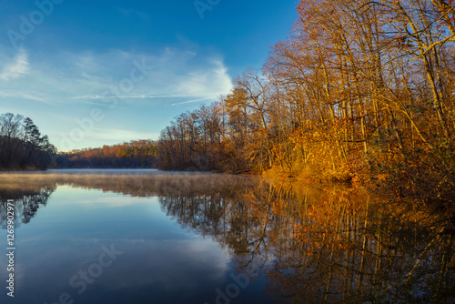 The morning sun casts a warm glow on the autumn trees along the banks of Chief McIntosh Lake in Indian Springs State Park in Georgia. The blue sky, clouds and trees reflect in the calm water.