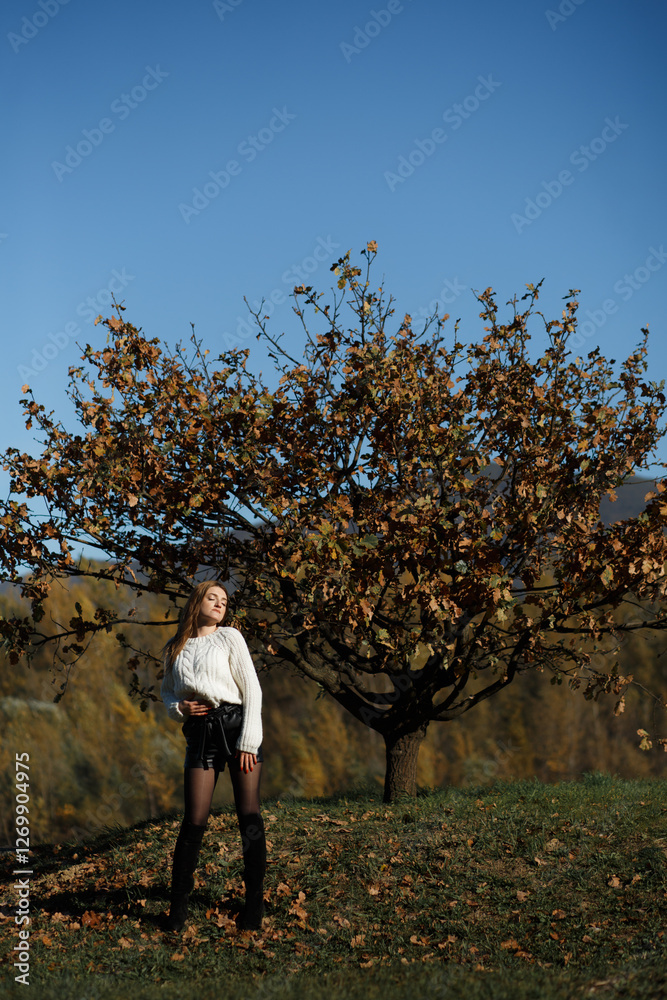 The girl stands with her eyes closed, placing one hand on her chest. Mountains and blue sky are visible in the background. The leaves on the tree have autumn colors such as brown and orange. The girl stands with her eyes closed, placing one hand on her chest. Mountains and blue sky are visible in the background. The leaves on the tree have autumn colors such as brown and orange.