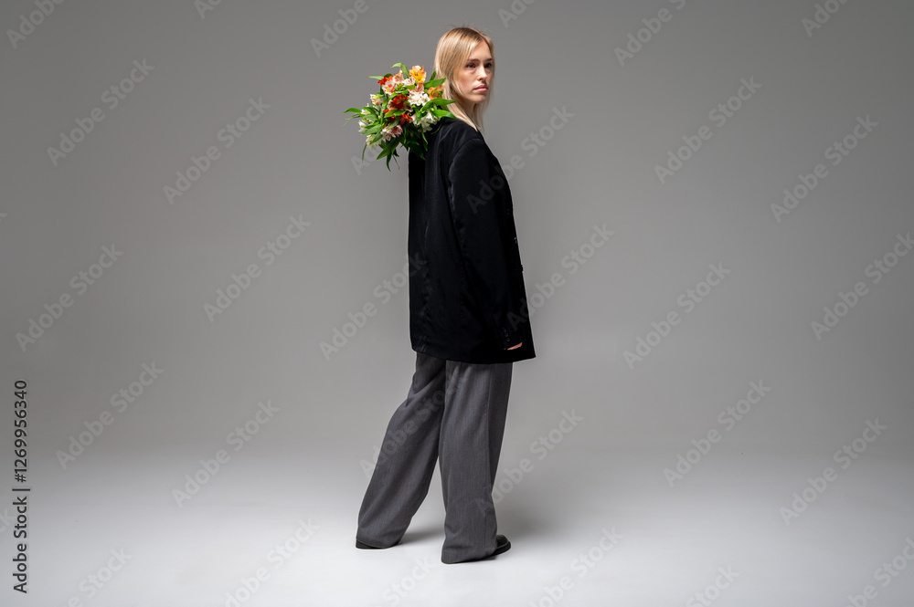 Stylish, young blonde girl in a black suit holds a bouquet of fresh flowers on her shoulder, view from the back, full length. Minimalistic studio portrait with a modern, elegant and fashionable look Stylish, young blonde girl in a black suit holds a bouquet of fresh flowers on her shoulder, view from the back, full length. Minimalistic studio portrait with a modern, elegant and fashionable look