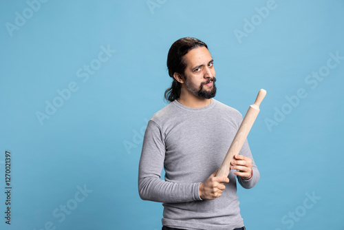 Furious serious person pointing a rolling pin at someone and starting a conflict, having an argument and being aggressive against blue background. Young man with rage and a violent reaction in studio.
