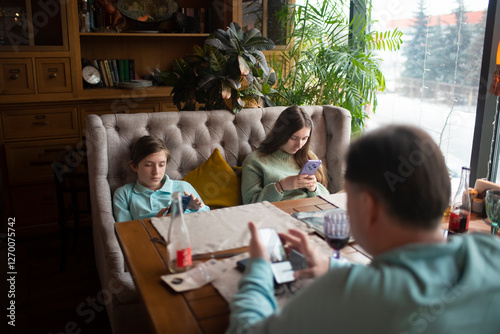 Family sitting at a table in a cafe, everyone looking at their smartphone. Children with their father waiting for their order in a restaurant, brother, sister and father surfing the Internet via mobil