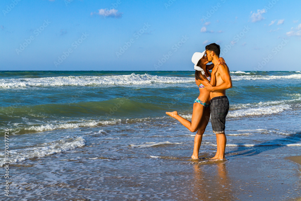 Happy young couple in bikini and shorts enjoying summer at the beach at dusk, breathing fresh air, kissing and teasing one another Happy young couple in bikini and shorts enjoying summer at the beach at dusk, breathing fresh air, kissing and teasing one another