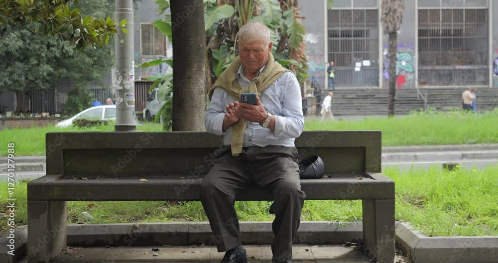 Old man rests on a bench in the city and uses his mobile phone