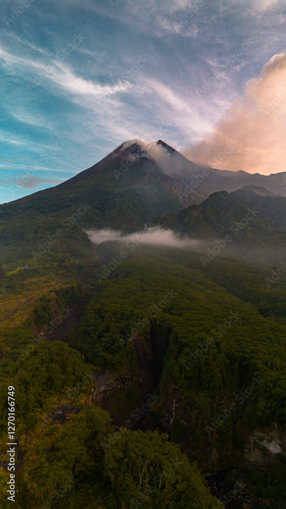 Fototapeta premium Portrait mode of a volcano view in the bright morning. Aerial photography of Mount Merapi in the morning with blue sky background and green hills in the foreground