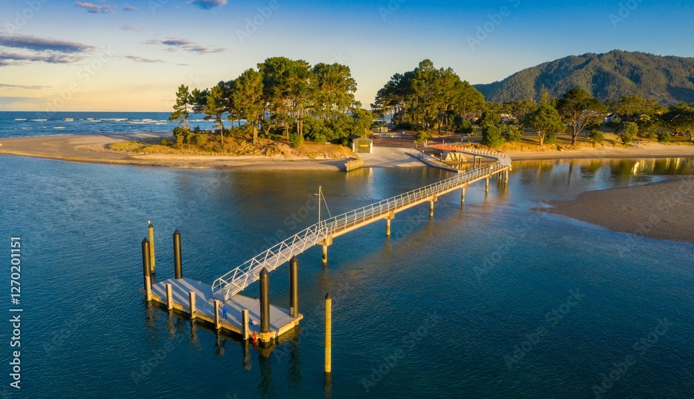 Fototapeta premium Aerial view of a scenic Pauanui boat ramp pier, with people using the walkway. Tranquil coastal scene at sunset, Coromandel Peninsula, New Zealand