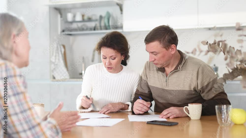 Thoughtful middle-aged man and woman sitting at the kitchen-table face to face with female agent demonstrating something on his laptop Thoughtful middle-aged man and woman sitting at the kitchen-table face to face with female agent demonstrating something on his laptop