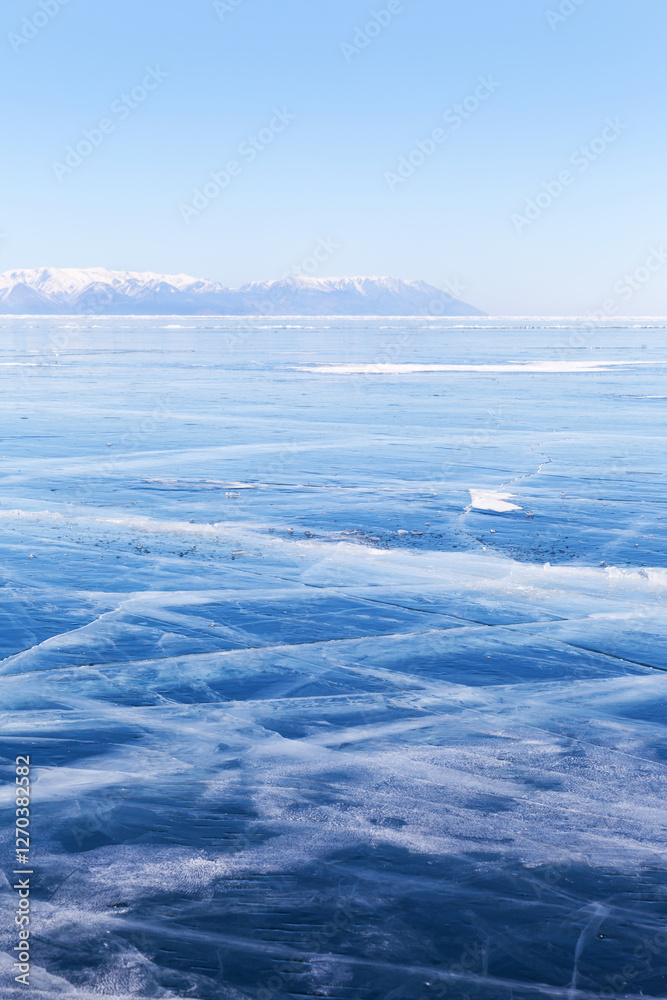 Obraz premium Typical winter landscape of frozen Baikal Lake. The endless fields of smooth blue ice with cracks and snowy mountains in the distance. Natural background. Winter travels and outdoors