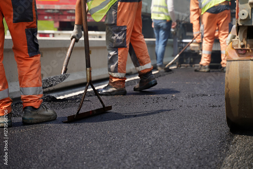 Fotografie Low-angle shot of multiple construction workers’ legs using tools to spread fresh asphalt next to a road barrier, highlighting teamwork and precision in road construction near barriers