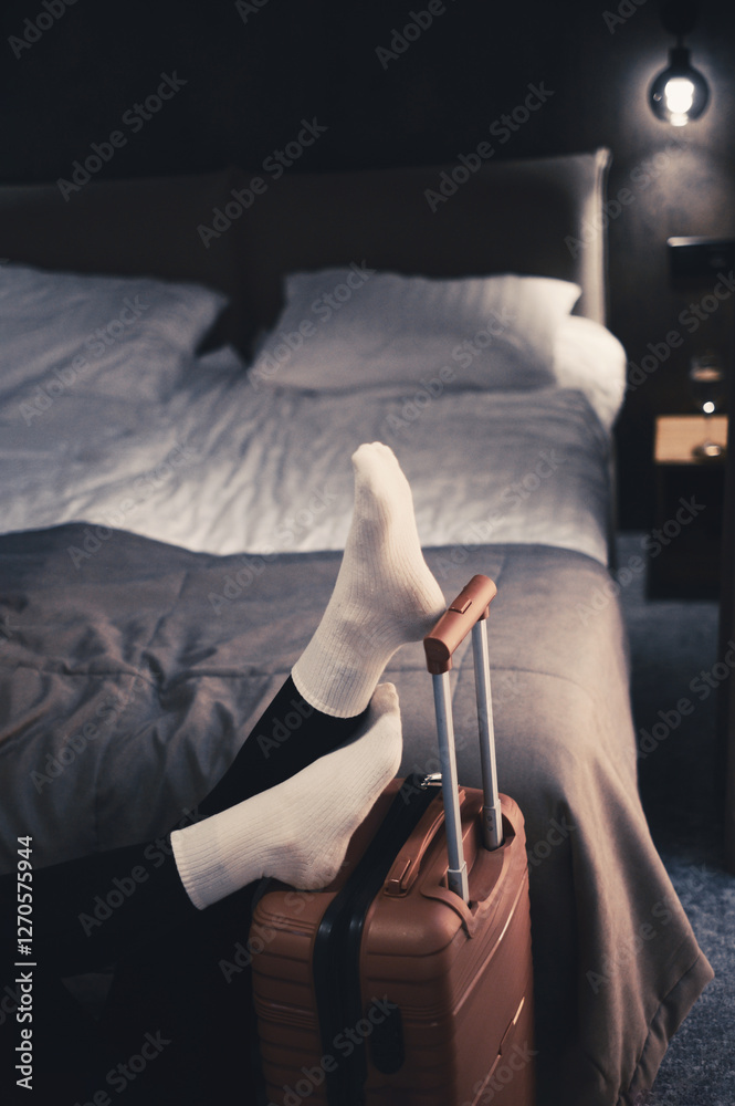 Obraz premium Woman lies on a bed in a hotel room with her legs on suitcase, showing a sense of solitude. Tired after long travel. Relaxation moment