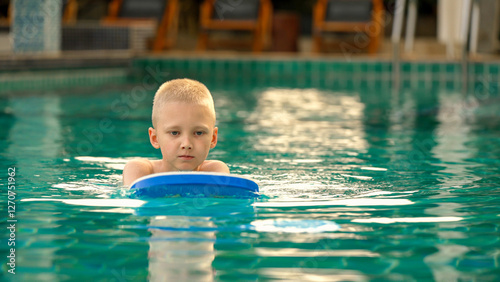 A young boy floats in a swimming pool on a swim board, concentrating as he practices his swimming skills. The relaxing environment features clear blue water and wooden lounge chairs nearby.