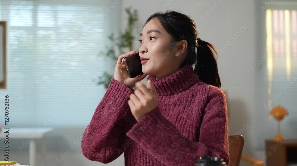 Comfortable and cozy, a young asian woman wearing a turtleneck sweater sits at her desk, engaged in a conversation on her mobile phone, gesturing as she speaks Comfortable and cozy, a young asian woman wearing a turtleneck sweater sits at her desk, engaged in a conversation on her mobile phone, gesturing as she speaks